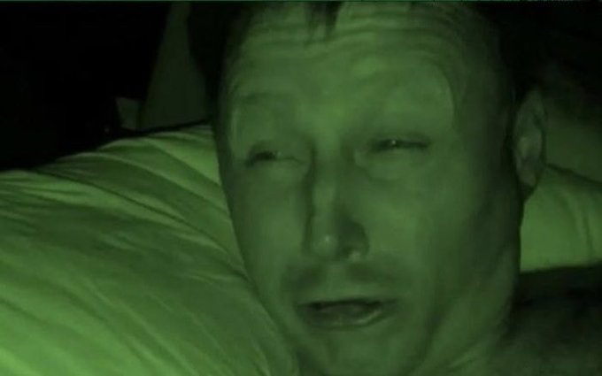 A night vision (green-tinted) close-up photo of a man with a mustache lying on a pillow, appearing exhausted or emotional with a distressed expression.