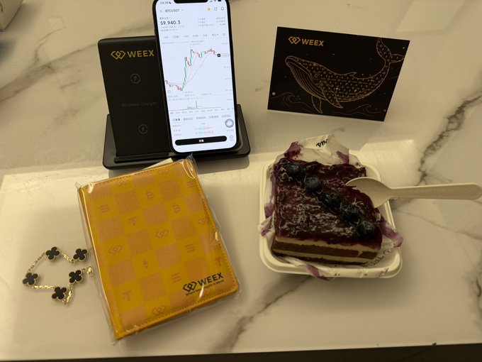 A flat lay photo on a marble surface featuring a smartphone displaying a $9,940.3 stock chart, a WEEX wireless charger, a WEEX whale-themed card, a yellow WEEX pouch, a clover bracelet, and a slice of blueberry cake.