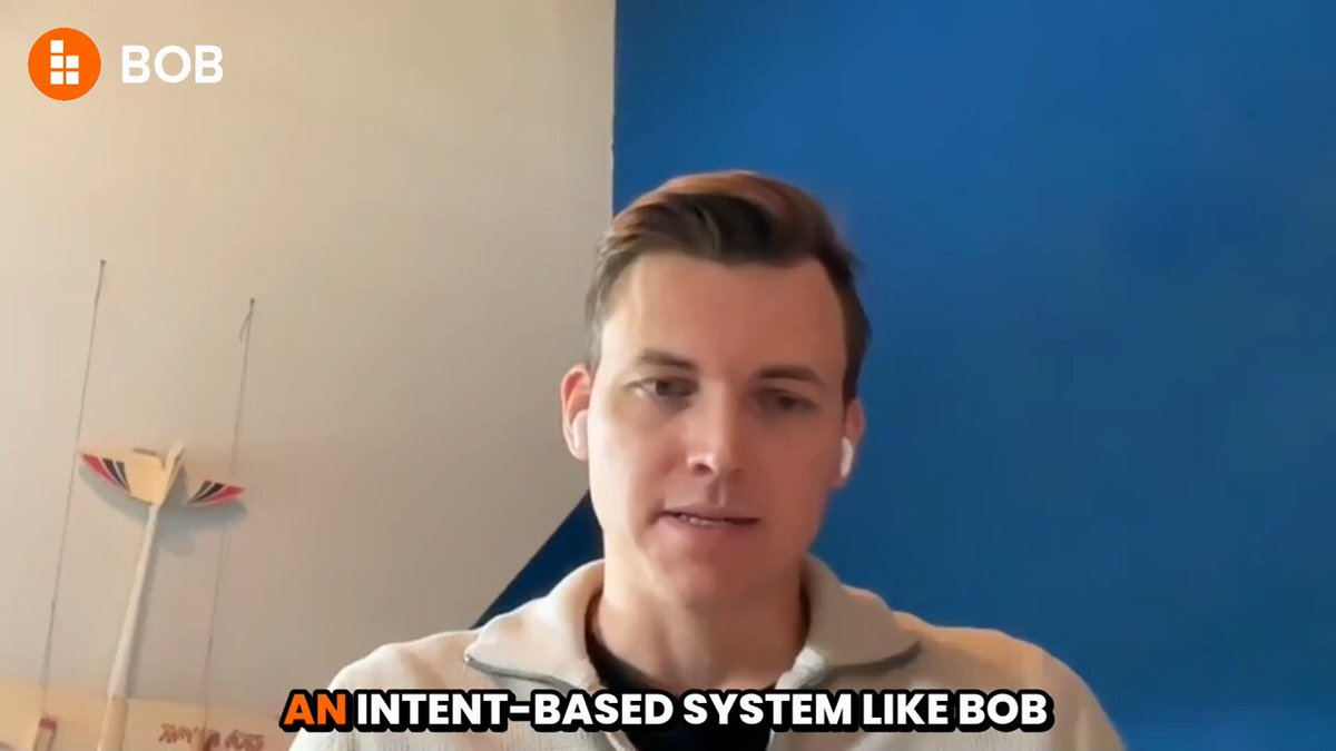 The image shows a man wearing a white shirt with black text that reads "An intent-based system like Bob". The background of the image features a blue wall with a white stripe running down its center, as well as an orange logo on the left side of the frame.