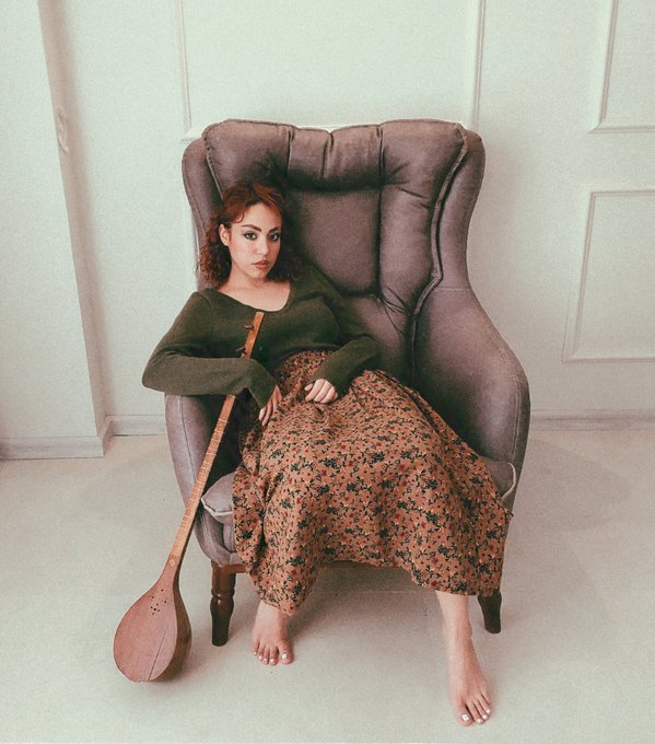 A woman with red hair sits barefoot in a large gray wingback chair, wearing a green sweater and floral skirt, holding a long-necked stringed instrument resting on the floor.