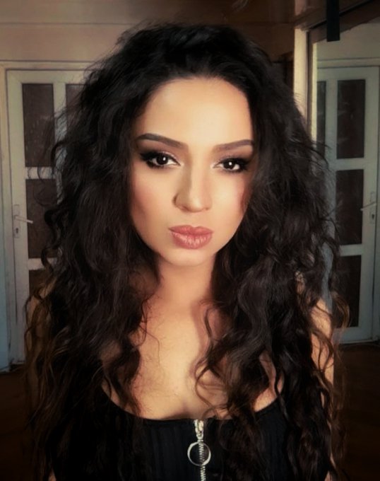 A woman with long, voluminous curly dark hair and heavy eye makeup poses in a black top.