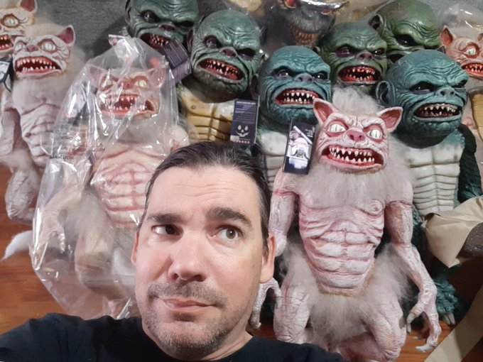 Man taking a selfie surrounded by an extensive collection of creepy creature masks and monster figurines, including pink and green ghoul designs.