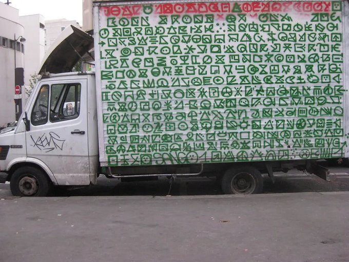 A white delivery truck with its cargo area covered in green graffiti tags and symbols parked on a street.