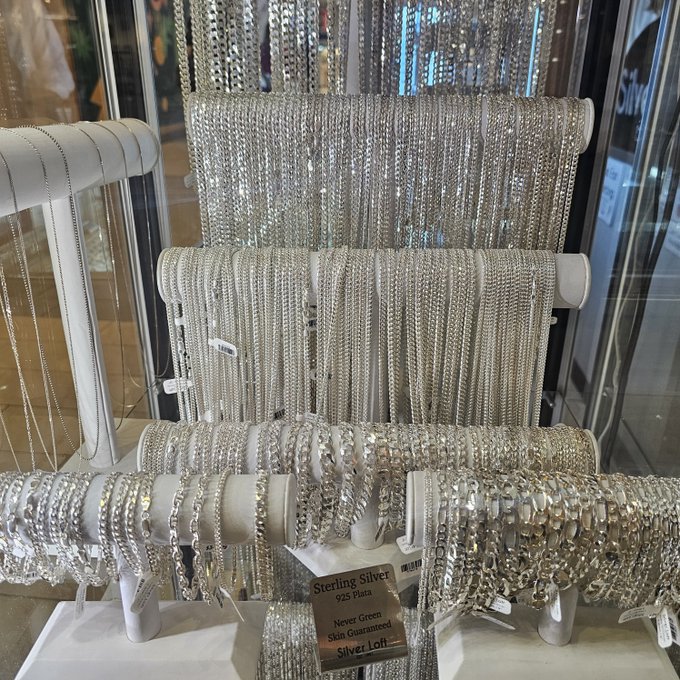 A display of sterling silver necklaces and chains at a jewelry store, with a sign reading 'Sterling Silver 925 Plata' and 'Never Green Skin Guaranteed Silver Lot'.