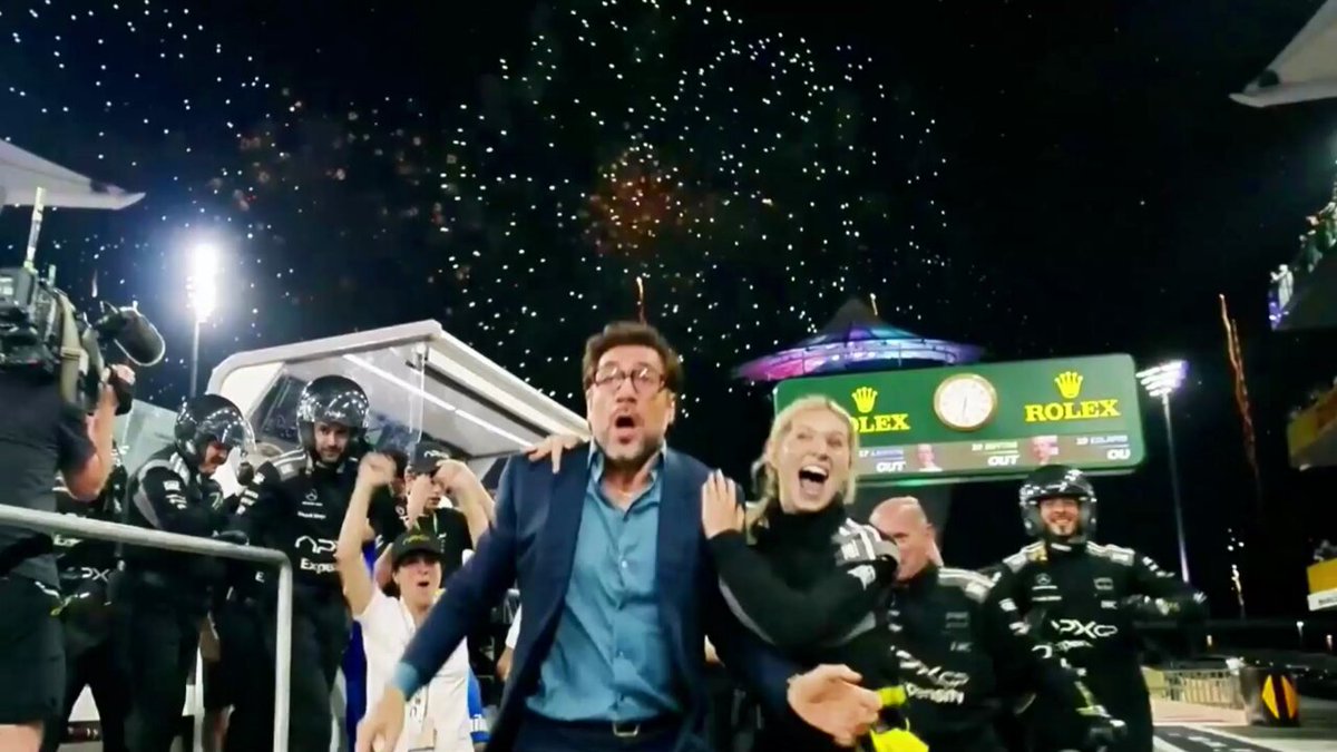 A sports event with people celebrating around a fireworks display, including a man wearing glasses holding his arm out for a high five while being photographed by multiple cameras.