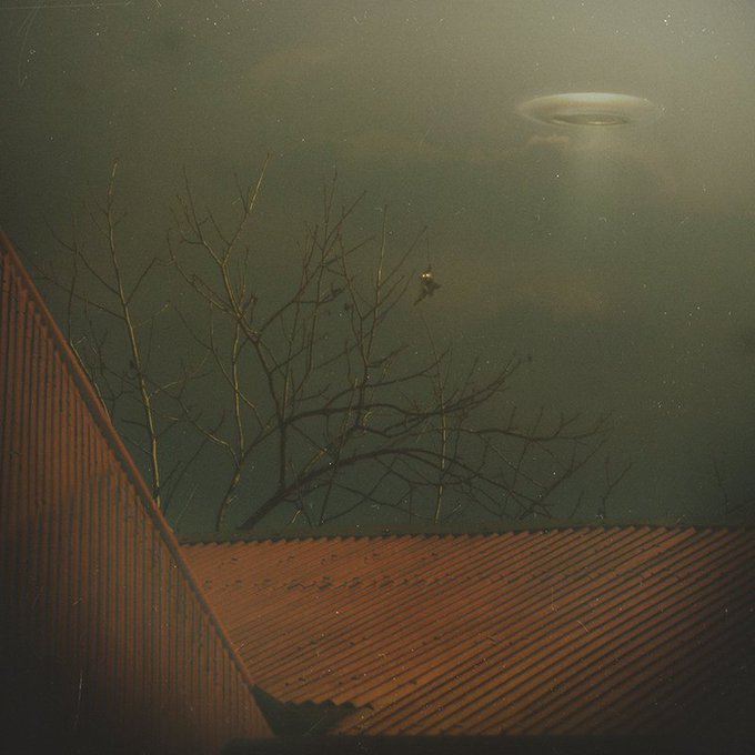 A nighttime photograph of a UFO hovering in the sky above a corrugated metal roof and bare tree branches.