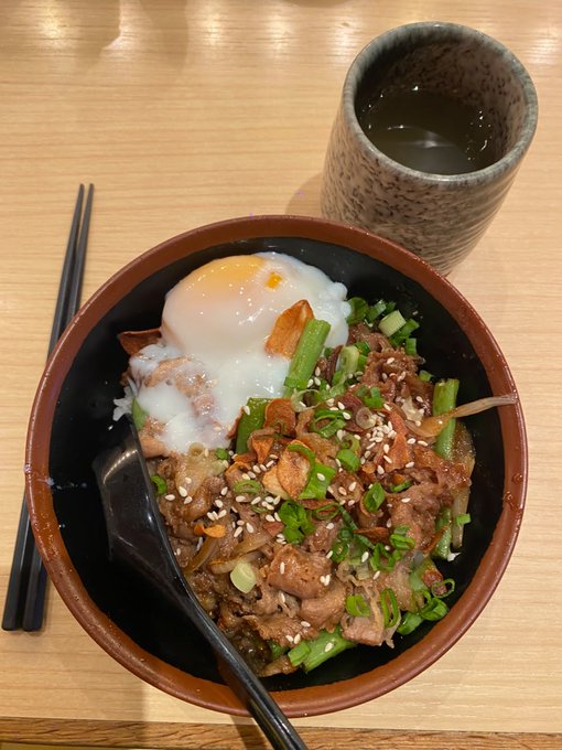 A bowl of rice topped with bulgogi-style beef, green onions, sesame seeds, fried garlic, and a soft-boiled egg, served with a spoon and chopsticks next to a cup of tea on a wooden table.