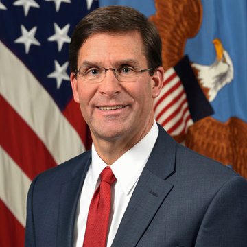 An official portrait of a smiling man in a navy suit and red tie, wearing glasses, posed in front of the American flag and a Department of Defense seal.