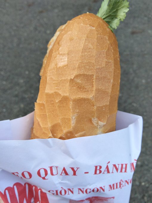 A bánh mì sandwich wrapped in paper printed with "QUAY - BÁNH" and "GIÒN NGON MIỆNG" sits on pavement, with fresh cilantro peeking from the top, accompanying a post about the poster's ongoing journey of eating bánh mì while building on X.
