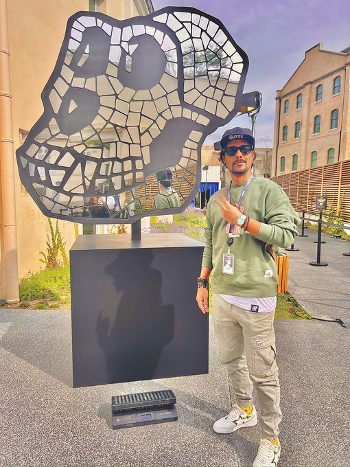 A man wearing sunglasses, a cap, and a green sweatshirt posing next to a metallic panther/puma sculpture outdoors.