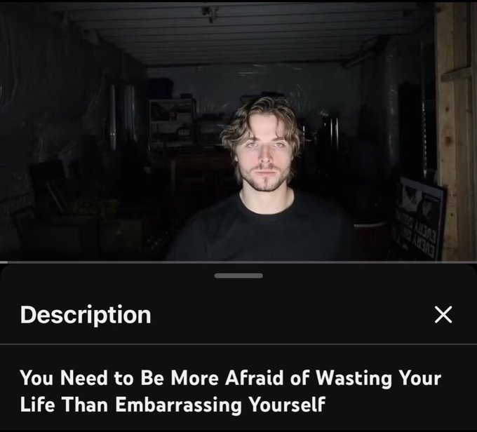 A man with wavy blonde hair and a goatee stands in a dimly lit garage, looking directly at the camera with a neutral expression. Below the photo, a "Description" overlay reads: "You Need to Be More Afraid of Wasting Your Life Than Embarrassing Yourself"—a motivational message about prioritizing meaningful action over fear of judgment, fitting the "Real" caption.