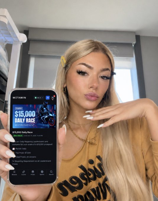 A blonde woman holds up her phone displaying the MetaWin app, which features a "$15,000 DAILY RACE" promotion with details about a "Daily Wagering Leaderboard" offering a $7,500 top prize and $20,000 in total prizes, directly illustrating the post's announcement about MetaWin's daily leaderboard rewards.
