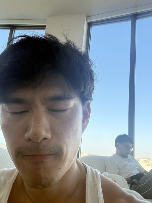 The image shows a man with dark hair wearing a white tank top, sitting next to another person who appears to be looking at him. They are both sitting indoors, possibly in front of a window or near a large glass surface like a mirror. The image also contains text that reads "bitcoin bounce now or i cut it off". This could potentially be an advertisement for Bitcoin or related products and services.