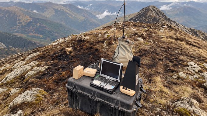 A rugged outdoor workstation is set up on a rocky mountain ridge, featuring an antenna, a rugged laptop, and small electronic devices housed in wooden enclosures mounted on a black hard case.