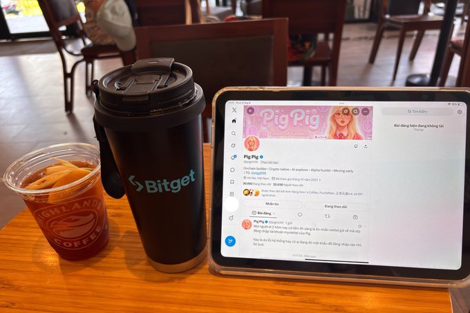 A coffee shop scene featuring a black Bitget branded tumbler and a clear iced coffee cup from Highlands Coffee next to a tablet displaying a Telegram chat with 'Pig Pig AI'.