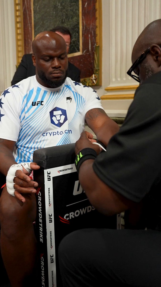 The image shows two men sitting next to each other, one of them wearing a UFC shirt. They are both holding boxing gloves, possibly preparing for a fight or practicing their skills. A third person can be seen standing behind the two men, likely observing or waiting for their turn in the ring.