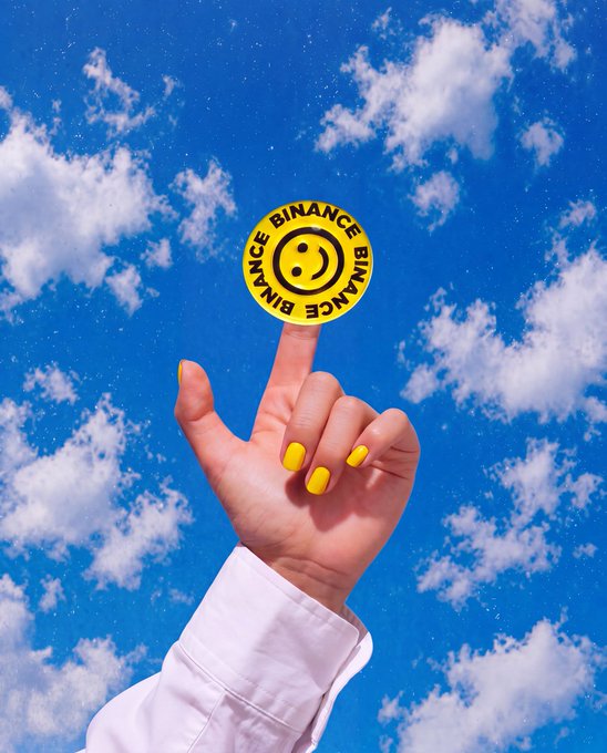 A hand with yellow nail polish balances a yellow Binance coin against a bright blue sky with scattered white clouds.