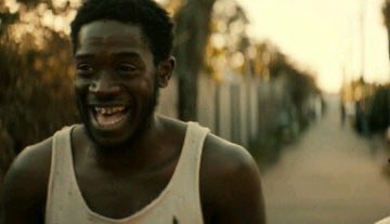 A still from a video showing a young Black man in a white tank top smiling joyfully outdoors at sunset.