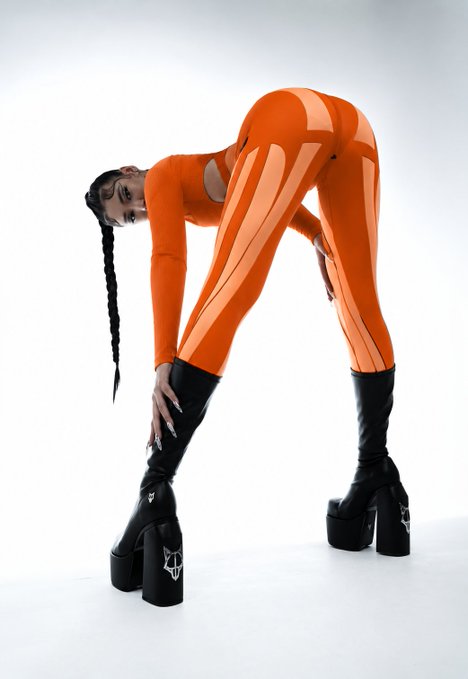 A model wearing a bright orange cutout bodysuit and towering black platform boots with logos, posing bent over against a white background.