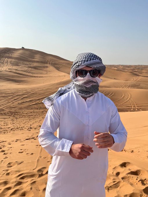 A man wearing traditional white Emirati attire and a black and white keffiyeh with dark sunglasses stands in a desert landscape with rolling sand dunes and a clear blue sky.