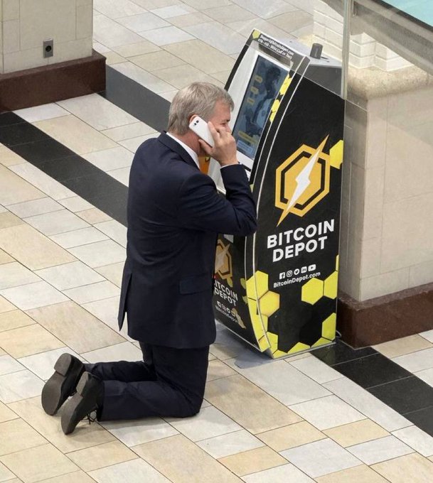 A man in a suit kneels on the tiled floor of a shopping mall while talking on a cell phone next to a yellow and black "BITCOIN DEPOT" ATM branded with bitcoindepot.com, symbolizing the growing mainstream financialization of Bitcoin that has led entities like Michael Saylor's Strategy to plan massive sales—up to $5 billion—of the asset to fund USD reserves, dividends, and share buybacks.