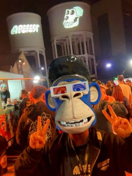 A person wearing a blue and white 3D-printed bear mask, making peace signs at night with illuminated tanks and a crowd in the background.