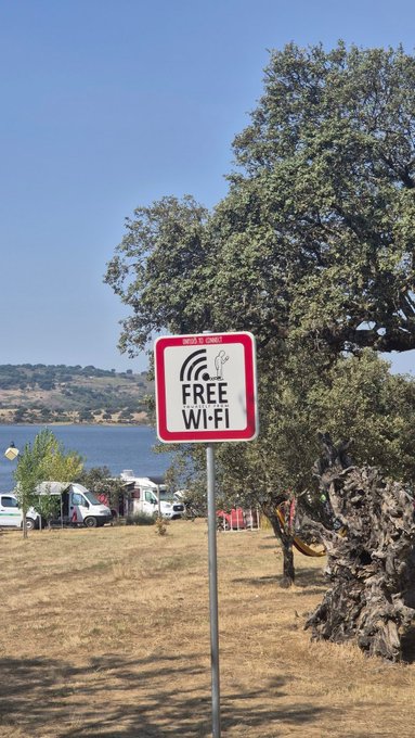 A red and white "FREE WI-FI" sign with a signal symbol stands in a dry, grassy campsite near a lake, surrounded by parked RVs and large oak trees. The post's reminder to "touch heart" playfully connects to the Wi-Fi sign, suggesting appreciation for the free connectivity at this scenic outdoor location.