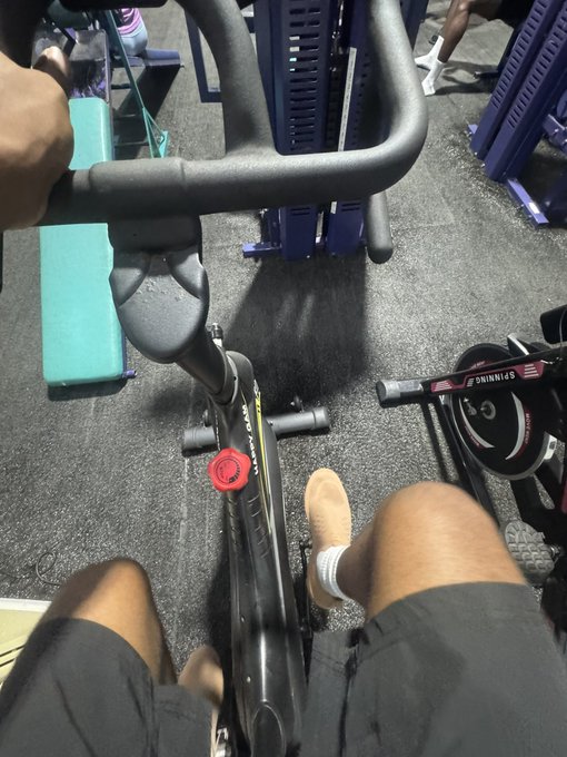 A first-person gym view shows someone seated on a stationary bike, with a “SHIMANO” weight plate and other exercise equipment nearby.