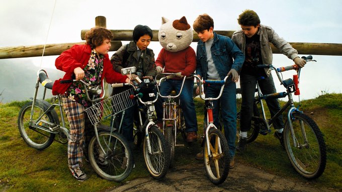 Five children, including one wearing a fox head mask, stand together with their BMX bicycles on a grassy hill.