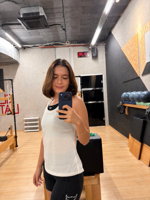 A woman takes a mirror selfie in a gym, wearing a white tank top and holding her phone, with a digital clock reading "06:13" visible on the wall behind her. The image captures a quiet, determined moment of early morning training, perfectly embodying her message of staying consistent and pushing forward one workout at a time.