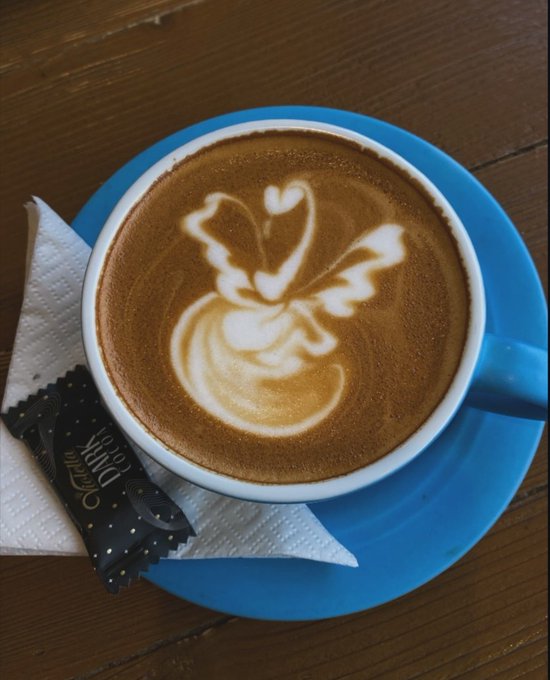 A blue ceramic cup of coffee served on a matching blue saucer with intricate latte art featuring a heart and leaf design, accompanied by a napkin and a 'Chocolat du Jour' chocolate.