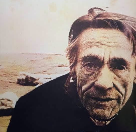 Portrait photograph of an elderly woman with weathered facial features on a coastal beach background with rocks visible.