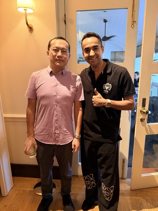 Two men pose for a photo inside a warmly lit restaurant or hotel lobby; one wears a pink short-sleeved button-up shirt while the other, in a black polo featuring the "OKX" logo on the chest, gives a thumbs-up. The image captures a meeting with Star Xu, founder and CEO of the cryptocurrency exchange OKX, reflecting the post's theme of connecting with a visionary crypto leader who remains focused on the product.
