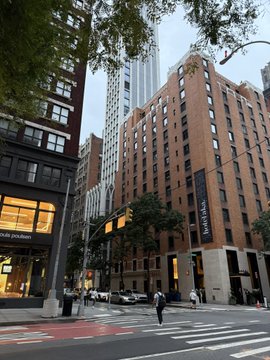 A street view of a Manhattan block featuring a tall red-brick building with a 'SHUBERT' banner, alongside retail storefronts including 'COACH' and 'LOUIS VUITTON.'