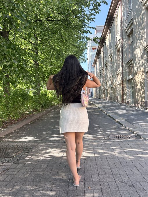 A woman with long dark hair walking down a tree-lined cobblestone street, wearing a black top and white skirt while carrying a pink purse.
