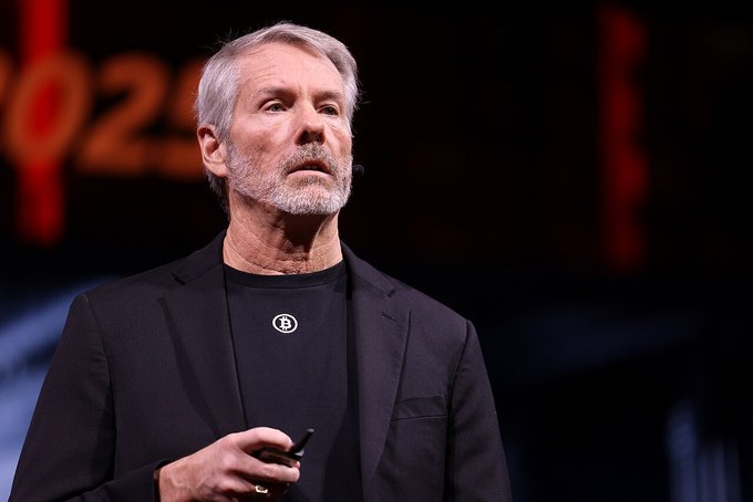 Photo of a gray-haired, bearded man in a black blazer speaking on stage, appearing to give a presentation.