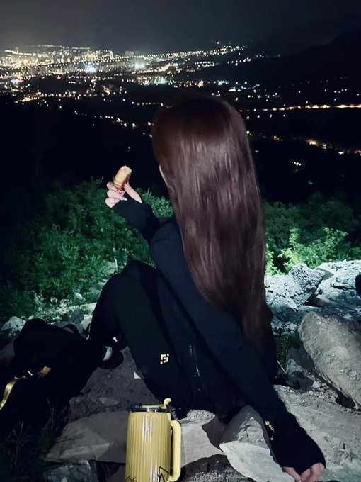 A person with long hair sits on a rocky hillside at night, gazing at a sprawling city skyline illuminated below, with a yellow lantern and a bottle beside them.