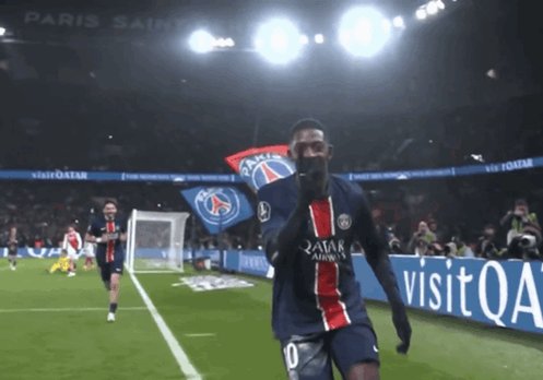 The image shows a soccer player wearing a blue and red jersey celebrating with his teammates after scoring a goal during a match at a stadium. The player has his hands up, possibly in a victory dance or expressing joy. In the background, there are other players on the field as well as spectators watching the game. A banner can be seen hanging from the ceiling of the stadium, and a soccer ball is visible near one of the players.