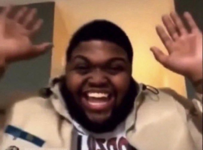 A blurry image of a man with a beard wearing a beige jacket over a t-shirt, smiling and holding his hands up near his face.