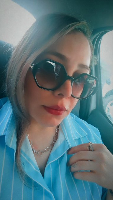 A woman wearing large sunglasses and a blue and white striped shirt takes a selfie inside a vehicle.