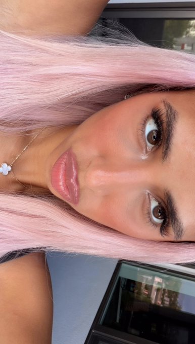 A close-up selfie of a woman with pastel pink hair, defined brows, glossy lips, and a four-leaf clover necklace, positioned sideways in the frame. The image visually complements the post's message of hopeful manifestation and optimism, pairing her confident, dreamy gaze with the caption's theme of chasing crypto goals and "retiring in 2026."