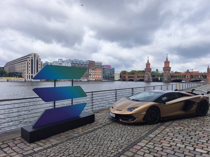 The image features a gold sports car parked next to a large stone wall, with a river running through the city behind them. In front of the car, there are three blue signs that appear to be part of an art installation or decoration for the Solana Summit event. The backdrop includes buildings and a bridge in the distance, creating a picturesque scene.