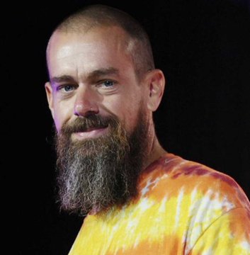 A close-up portrait of a smiling, bald man with a thick brown beard wearing a yellow and orange tie-dye shirt against a black background.