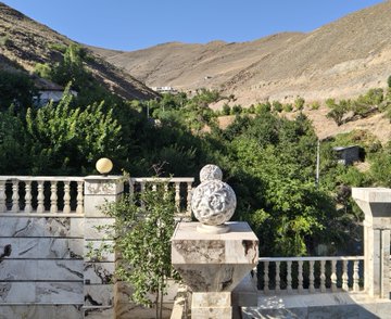 A scenic outdoor view featuring a marble balustrade with decorative spheres overlooking a valley of green trees and dry mountains under a clear blue sky.