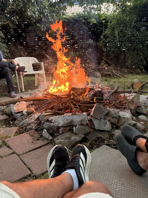 A small outdoor fire burns in a makeshift stone-and-branch pit, surrounded by seated people whose legs and shoes are visible in the foreground.