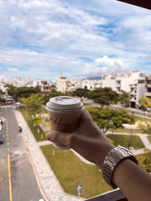 A hand holding a paper coffee cup with 'Aimee' printed on it, with a wristwatch visible, set against a backdrop of a city street and buildings under a cloudy sky.