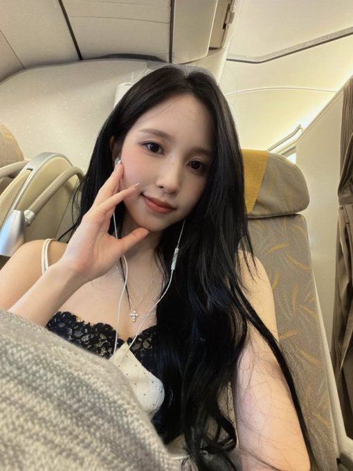 A selfie of a young woman with long dark hair wearing earphones and a black lace top, sitting in what appears to be an airplane seat.