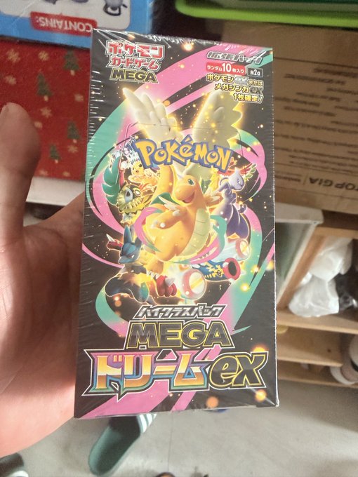 A hand holds a sealed Pokémon Trading Card Game MEGA Dream ex booster box featuring Charizard, Eevee, and Lucario artwork on the packaging.