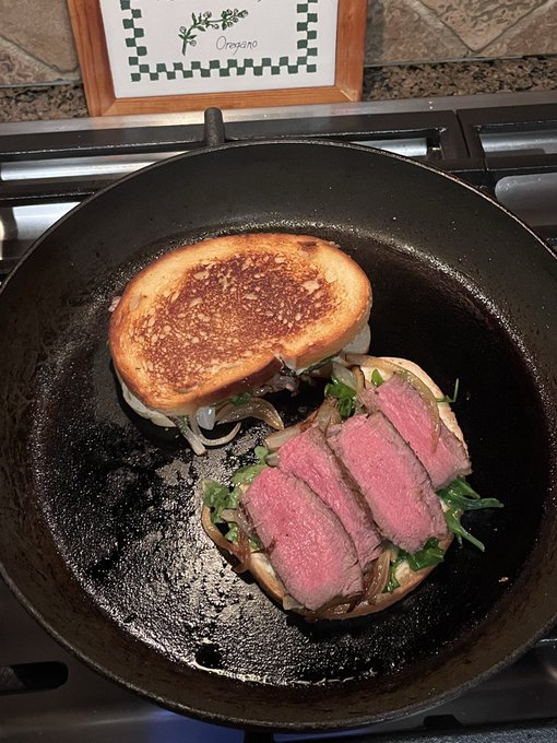 A cast iron skillet holds an open-faced steak sandwich with golden toasted bread, caramelized onions, arugula, and slices of medium-rare steak, prepared again after a first satisfying attempt. A framed "Oregaro" sign sits in the background above the stove.