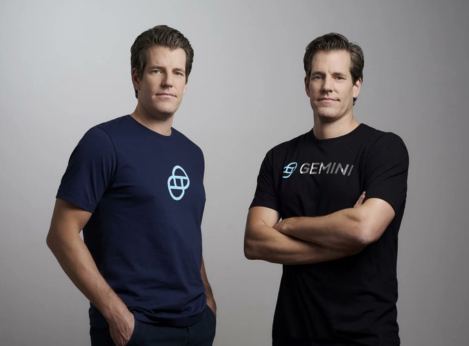 Two men, appearing to be twins, pose against a plain gray background—one wearing a navy blue t-shirt with a light blue Gemini-style logo, and the other wearing a black t-shirt featuring the "GEMINI" wordmark—visually representing the Gemini exchange brand associated with the announcement of native XRP Ledger deposit and withdrawal support in Singapore.